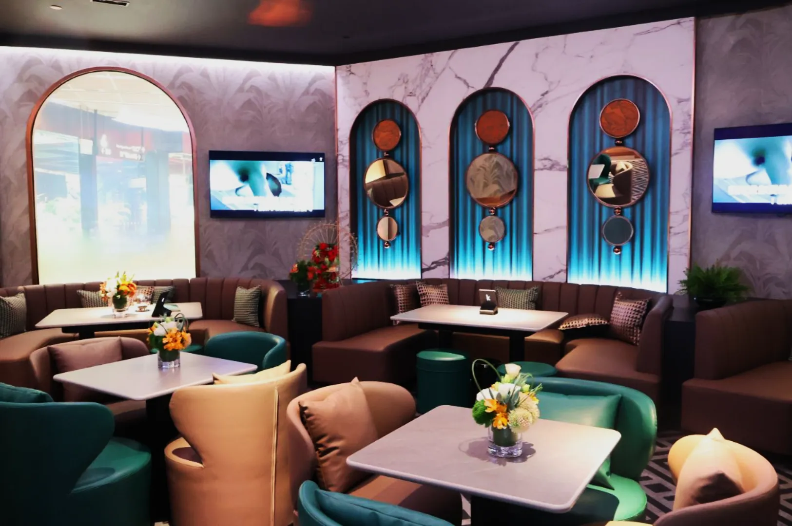 Chic lounge with plush beige and teal seating, marble accents, round mirrors, and floral arrangements. Relaxing ambiance with muted lighting and wall art.