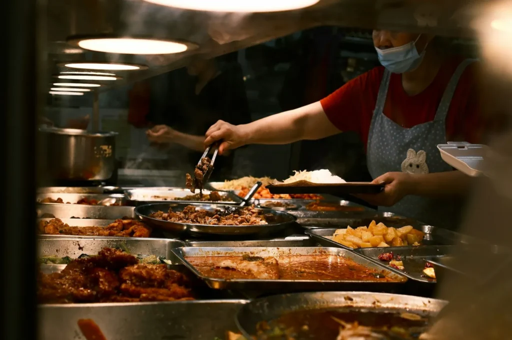 A person wearing a mask and apron serves food with tongs from metal trays in a dimly lit cafeteria. Various dishes, including rice and meats, are on display.