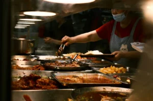 A person wearing a mask and apron serves food with tongs from metal trays in a dimly lit cafeteria. Various dishes, including rice and meats, are on display.