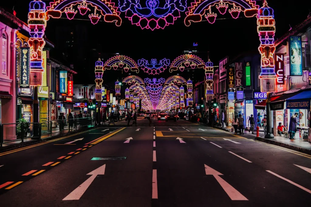 A vibrant city street at night, lit by intricate, colorful archway lights stretching into the distance. Neon signs and shops line both sides, creating a festive mood.