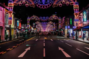 A vibrant city street at night, lit by intricate, colorful archway lights stretching into the distance. Neon signs and shops line both sides, creating a festive mood.