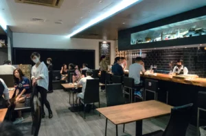 Modern restaurant with dim lighting, featuring patrons seated at black tables and barstools. A masked server moves between tables, creating a cozy ambiance.