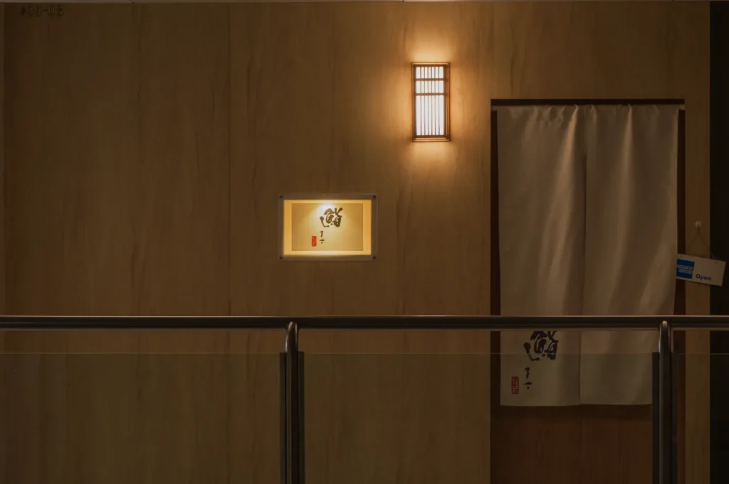 Dimly lit wooden interior with a glowing wall light above a framed artwork. A beige curtain hangs beside it, creating a serene, minimalist ambiance.