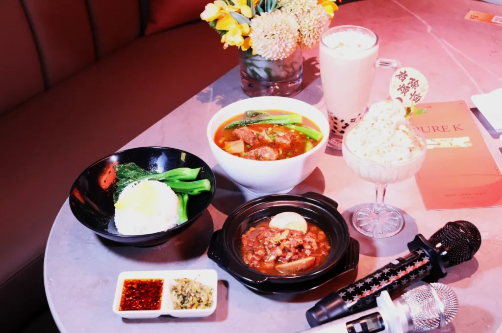 A table setting of Asian cuisine includes a bowl of rice with greens, a bowl of soup, and a clay pot dish. Nearby are sauces, a bubble tea, and a dessert. A vase of flowers and two microphones add a lively, inviting atmosphere.