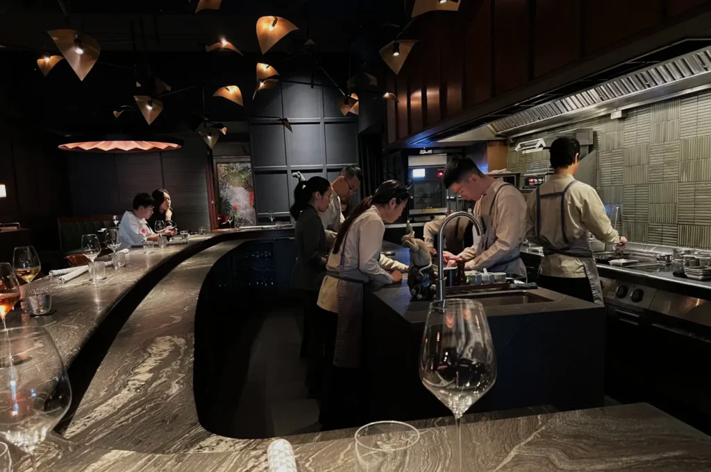 Dimly lit upscale restaurant with chefs preparing food in an open kitchen. Guests sit at a curved marble bar, wine glasses in the foreground.