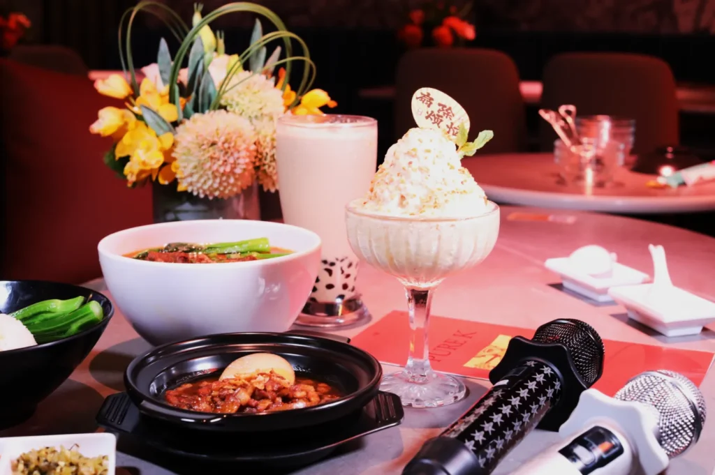 A table with a varied meal, including a bowl of soup, a dessert in a glass, bubble tea, and a floral centerpiece. Two microphones suggest a karaoke setting.