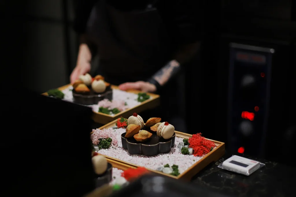 A chef presents two trays of artisanal desserts arranged with precision on a bed of white and red flakes, accented by small greens, in a dimly lit setting.