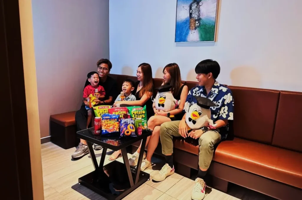 A group of five smiling people, including two children and three adults, sit on a brown bench in a cozy room. Snacks like chips and candy are on the table.