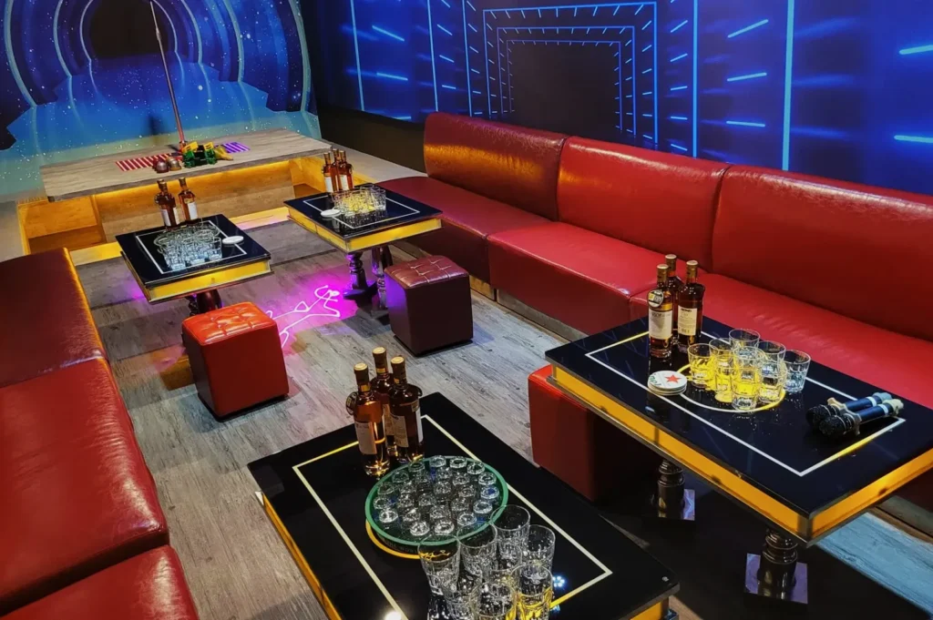 Spacious karaoke room with red leather seating, futuristic neon wall panels, and tables set with bottles, glasses, and snacks, creating a lively atmosphere.