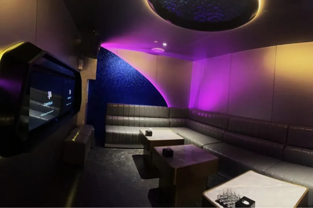 A sleek, modern karaoke room with a large screen on the wall, gray plush seating, purple ambient lighting, and tables creating an inviting atmosphere.