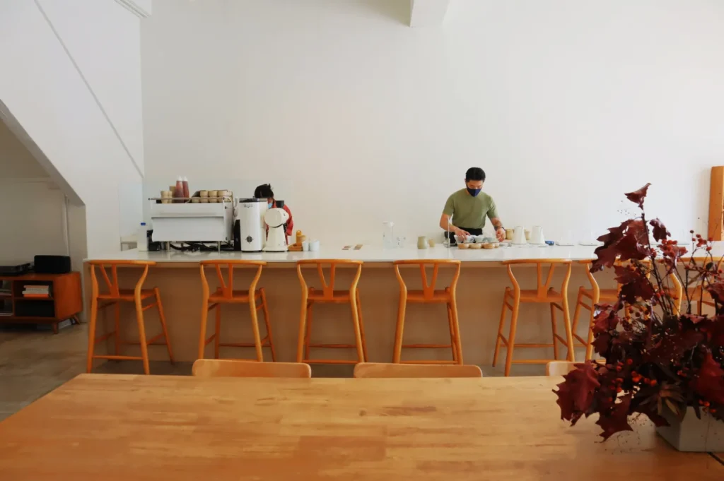 A barista wearing a mask prepares drinks behind a modern café counter with wooden stools. A table with autumnal flowers is in the foreground.