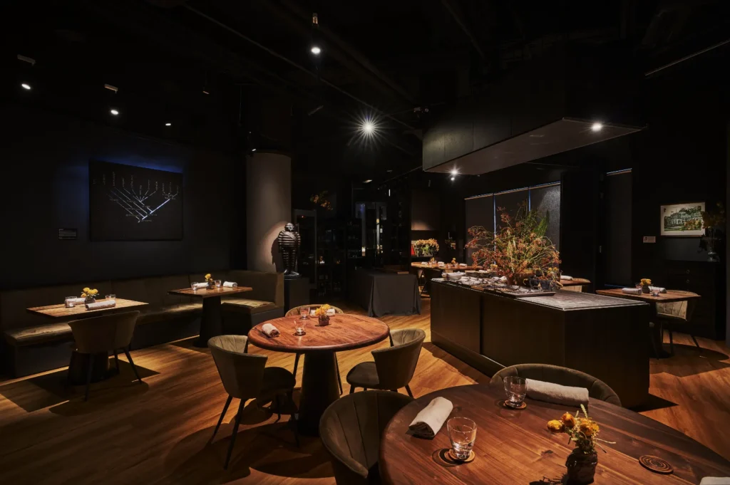 Cozy restaurant interior with round wooden tables, plush chairs, and dim lighting. Elegant decor includes wall art and floral arrangements, creating a warm, inviting atmosphere.