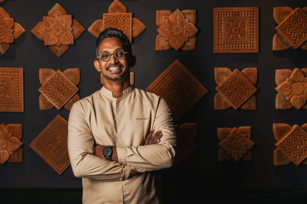 Smiling man with glasses and a light-colored chef jacket stands with arms crossed. Background features decorative wooden carvings in geometric patterns.