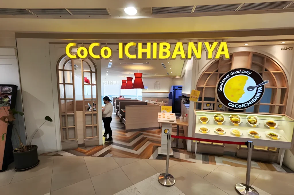 Entrance of CoCo ICHIBANYA restaurant with a warm, inviting interior. Yellow signage, display of curry dishes, and a person standing inside.