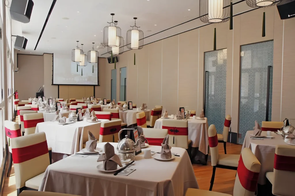 Elegant dining room with neatly arranged tables, white tablecloths, and red-accented chairs. Soft lighting from modern chandeliers creates a warm, inviting atmosphere.