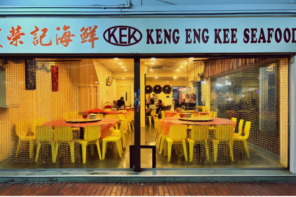 Front view of Keng Eng Kee Seafood restaurant with warm lighting. Yellow chairs and round tables are visible through the glass façade, creating a cozy, welcoming atmosphere.