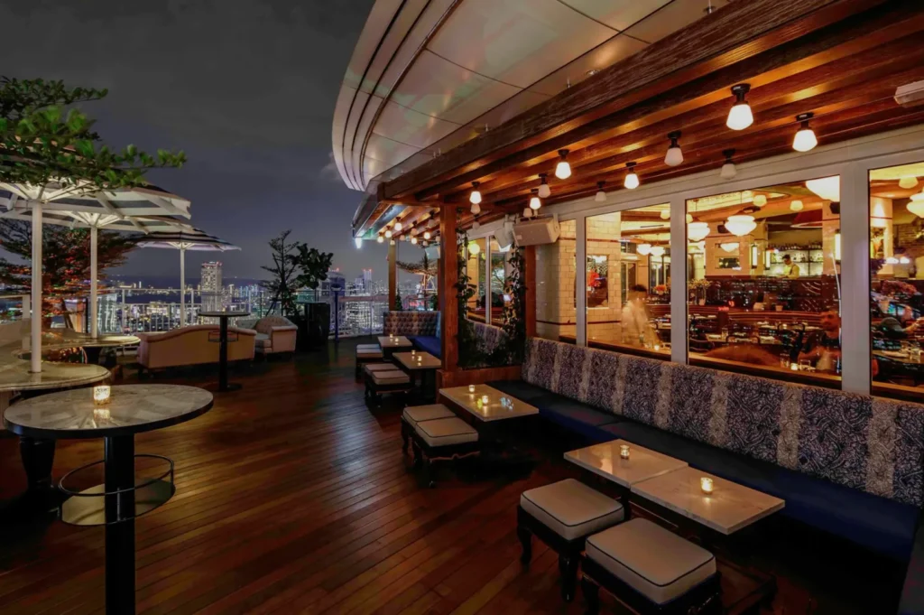Elegant rooftop bar at night, featuring cozy seating with cushions, dim lights, and city skyline views. Inviting, warm ambiance with wooden floors.