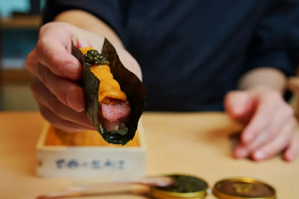 A hand presenting a sushi roll wrapped in seaweed, topped with uni and caviar. The background is a blurred dining setting, emphasizing a gourmet experience.
