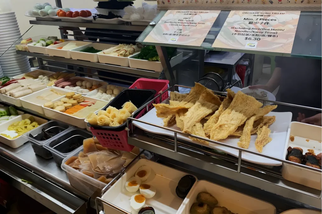 Self-service buffet counter with assorted Asian ingredients, including tofu, vegetables, and hard-boiled eggs. Signs display pricing and dish options.