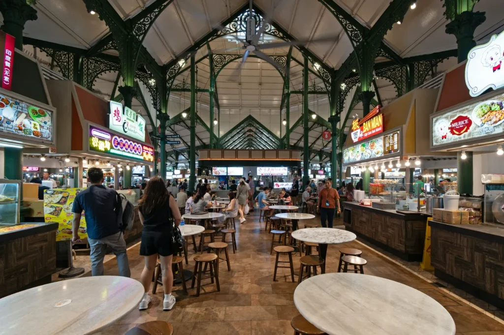 Bustling indoor food market with high ceilings, ornate green pillars, and various food stalls. People are walking and seated at round tables, creating a lively, inviting atmosphere.
