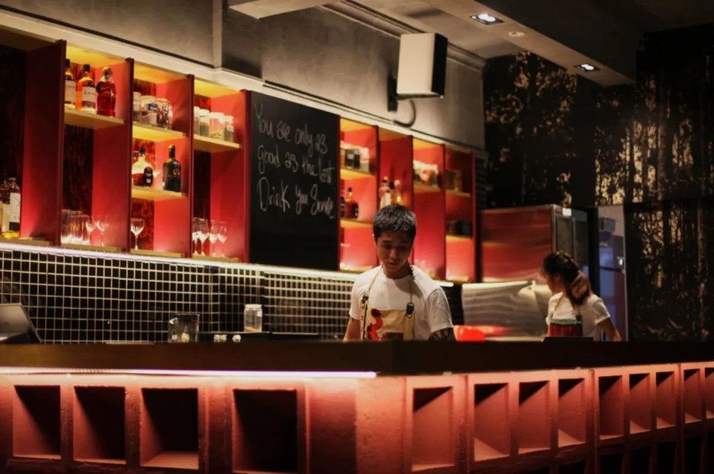 A dimly lit modern bar with two bartenders in white shirts. Shelves with bottles and glasses line the wall. A chalkboard reads, "Drink you smile."
