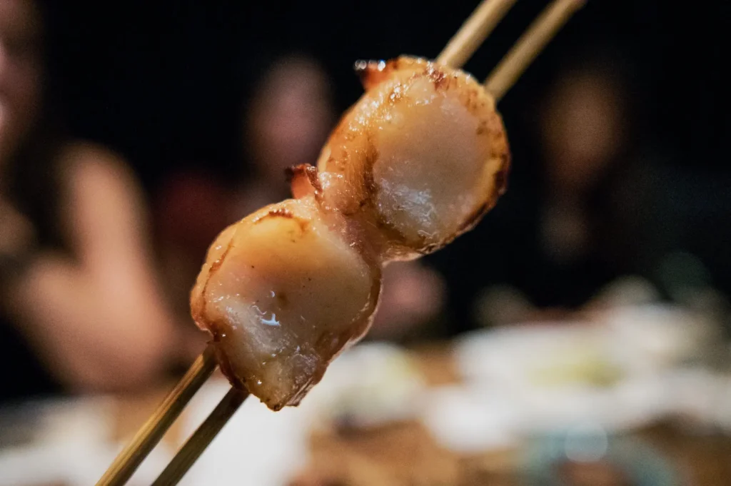 Close-up of two grilled scallops on skewers, glistening with a slight char. Background shows a dimly lit dining setting with blurred figures.