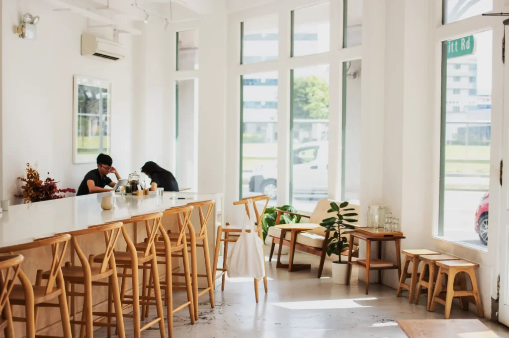 Bright, minimalistic cafe with wooden chairs, large windows, and natural light. Two people sit at a table, creating a calm and cozy atmosphere.