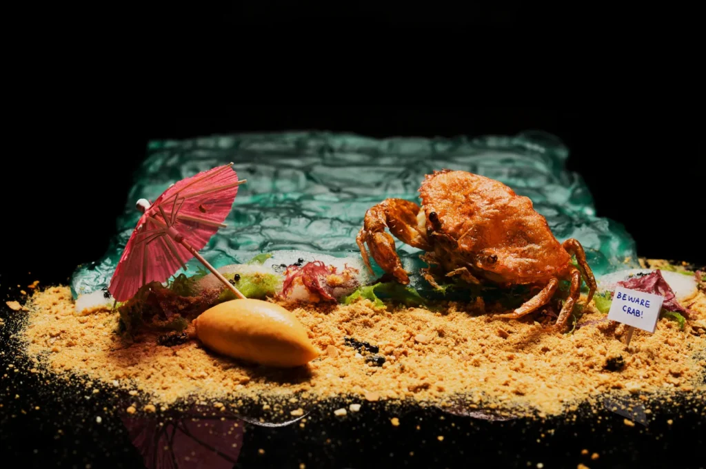 A whimsical food scene depicts a fried crab on a sandy surface with breadcrumbs, a pink cocktail umbrella, a pastry, and a "Beware Crab!" sign.