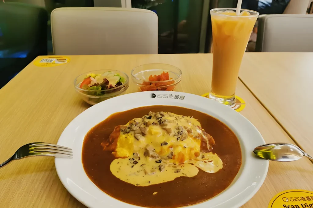 A plate of creamy mushroom sauce omurice sits on a table with curry and rice, accompanied by a salad, kimchi, and an iced latte, conveying a cozy, inviting meal scene.