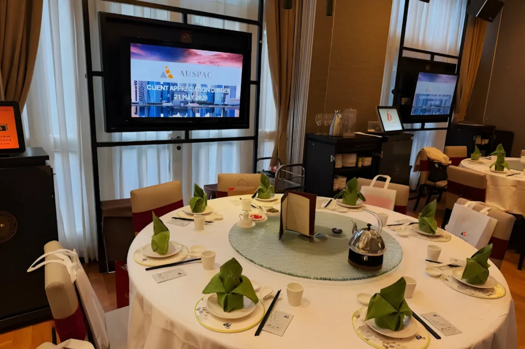 A round banquet table is elegantly set with white tablecloth, green napkins folded into lotus shapes, and tableware. A large screen in the background reads "Client Appreciation Dinner." The ambiance is formal and inviting.