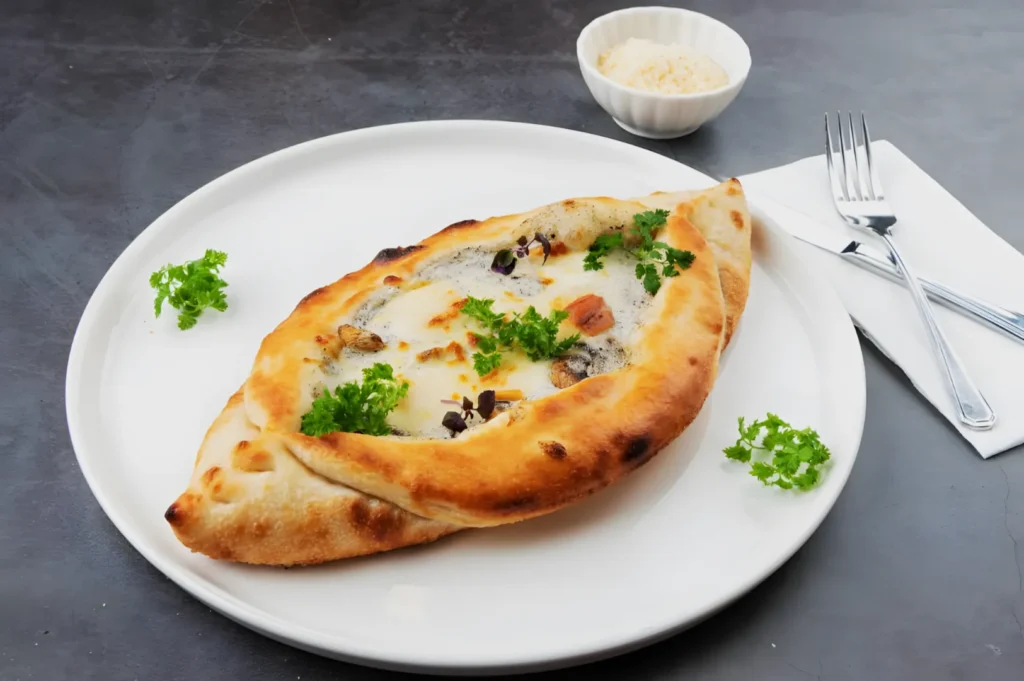 A baked flatbread topped with cheese, herbs, and vegetables is on a white plate with parsley garnish. A fork and knife lie on a napkin nearby, and a small bowl of grated cheese is in the background.