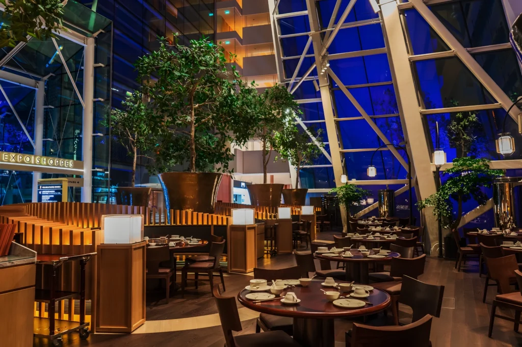 Modern restaurant with warm lighting, lush green plants, wooden furniture, and a large glass window revealing a blue-lit evening skyline.