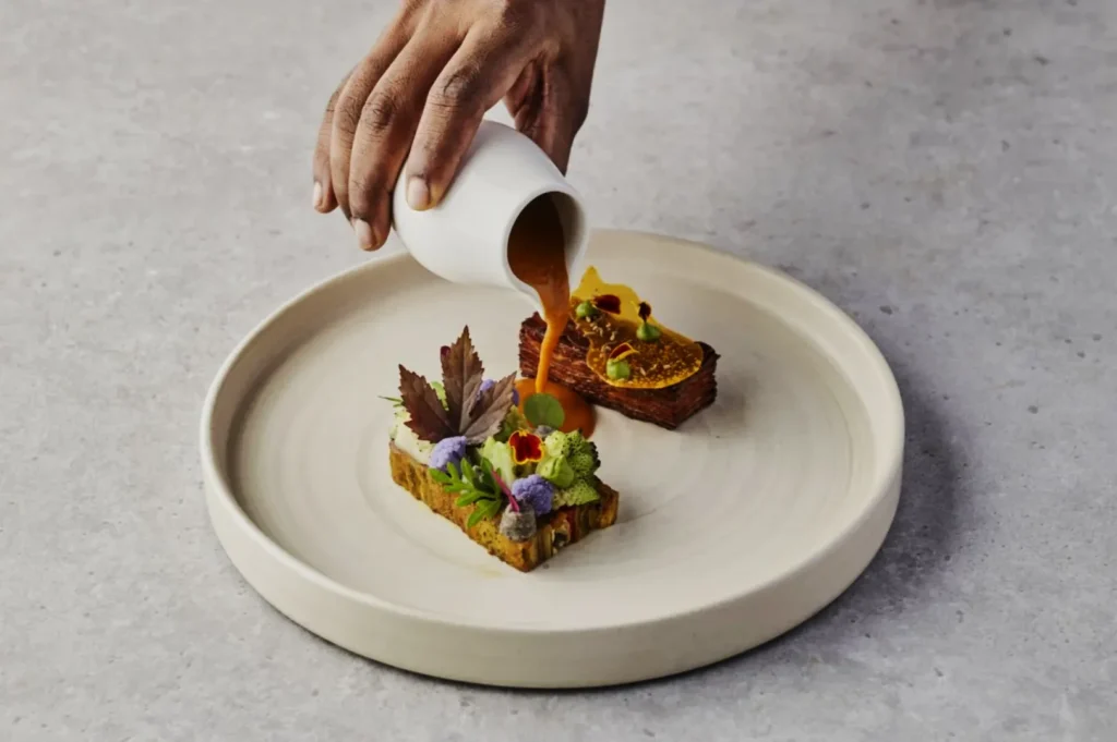 A hand pours a rich, brown sauce from a white jug onto an artfully plated dish. The dish features colorful garnishes on a light ceramic plate.