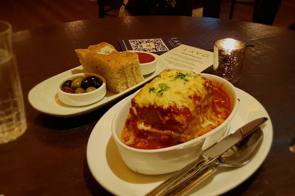 A close-up of a hearty lasagna topped with melted cheese and herbs, served with a side of bread and olives. A candle adds warmth to the cozy ambiance.