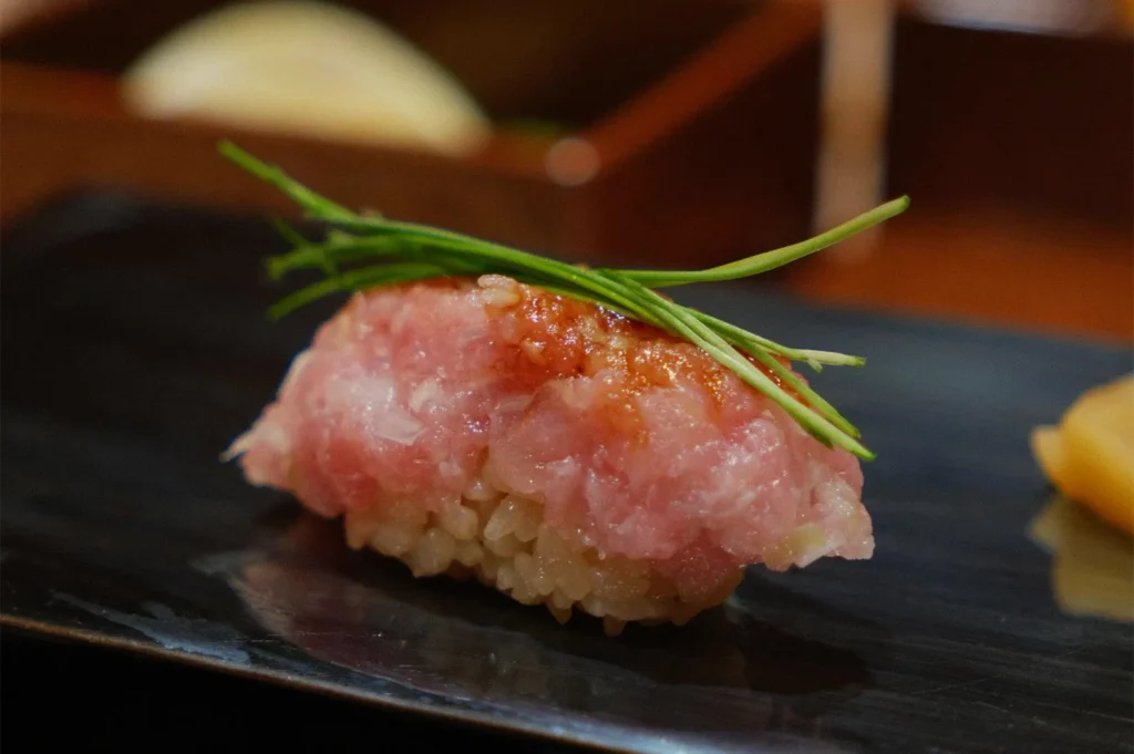 A piece of sushi with minced raw tuna on rice, topped with green chives, sits on a dark rectangular plate. It exudes elegance and freshness.
