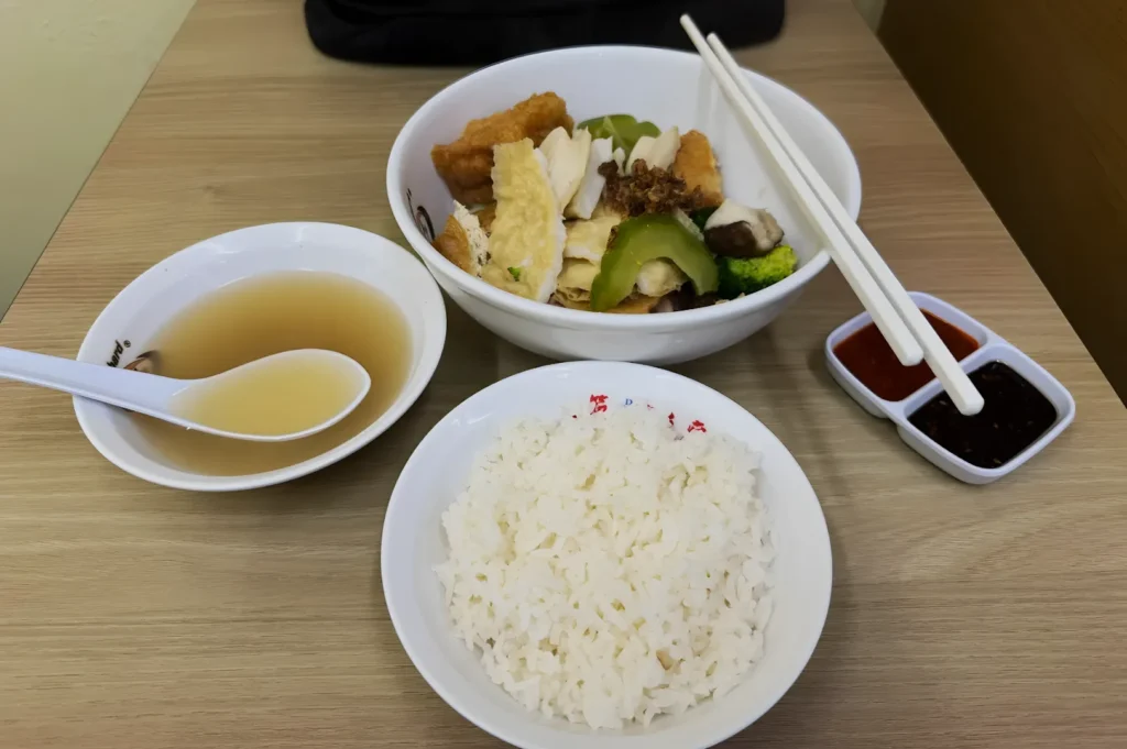 A bowl of white rice, a bowl with tofu, mushrooms, broccoli, and chicken, and a bowl of clear soup on a wooden table. Chopsticks and dipping sauces included.