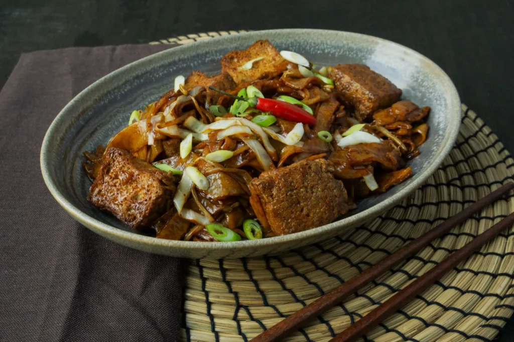 A rustic bowl of stir-fried tofu with green onions, cabbage, and red chili peppers on a woven mat, accompanied by wooden chopsticks.