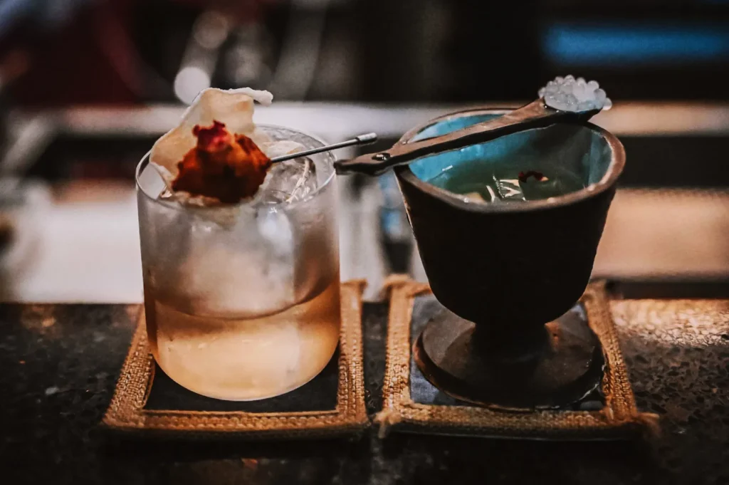 A cocktail with ice and a garnish sits on a coaster next to a small cup with a spoon of salt. The scene conveys an artisanal, refined vibe.