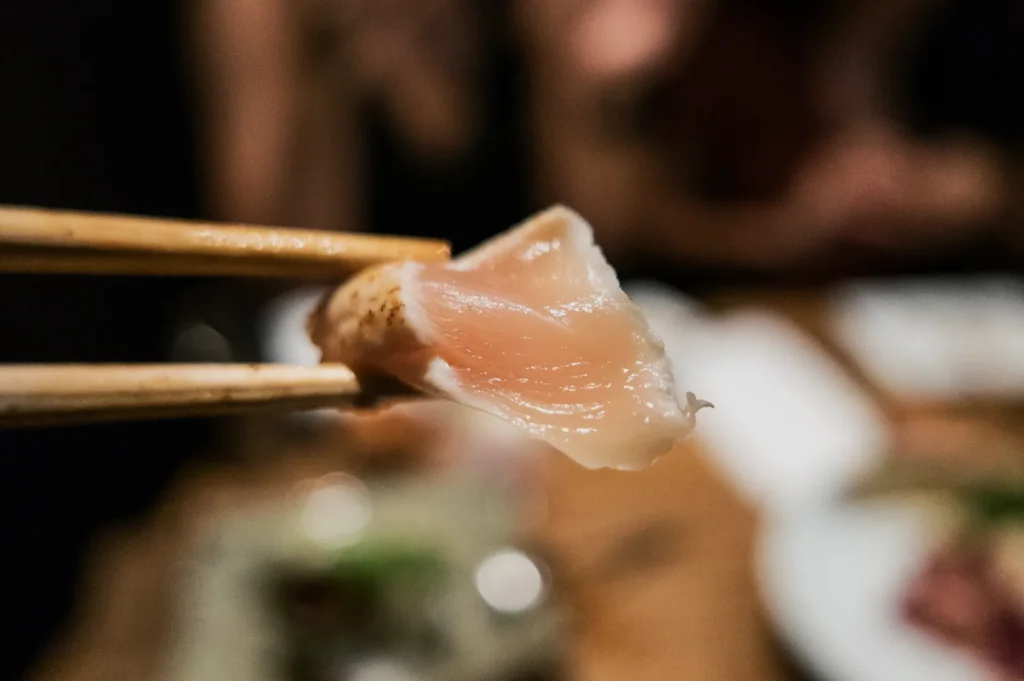 Close-up of chopsticks holding a piece of fresh, pink sushi, with a blurred restaurant setting in the background, conveying a casual dining experience.