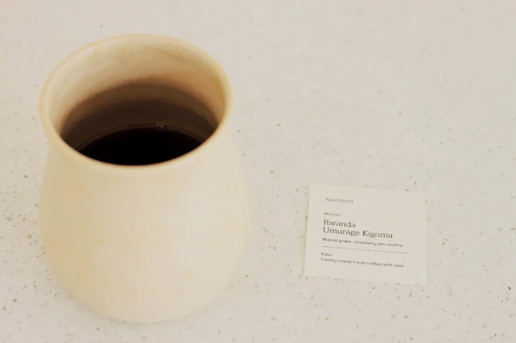 A beige ceramic cup filled with dark coffee sits on a light speckled surface. Beside it, a note reads “Rwanda Umurage Kigoma,” referencing coffee notes.