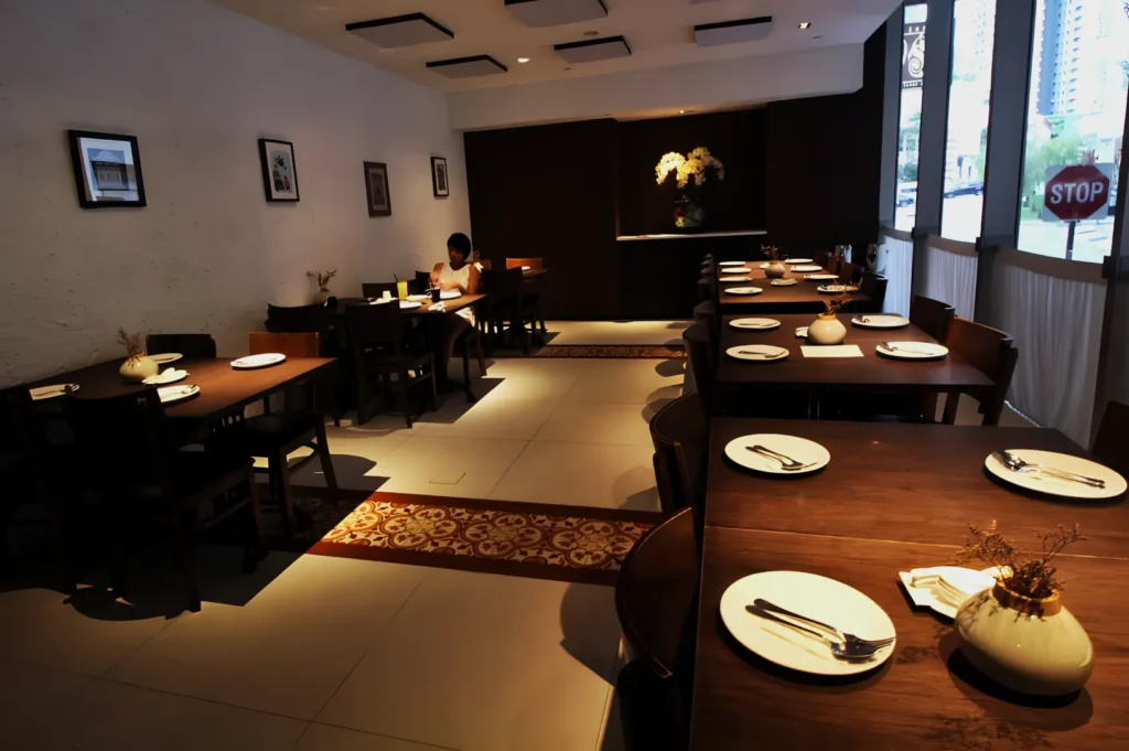 Cozy restaurant interior with wooden tables set for dining, featuring white plates and utensils. A lone person sits by the wall under soft lighting.