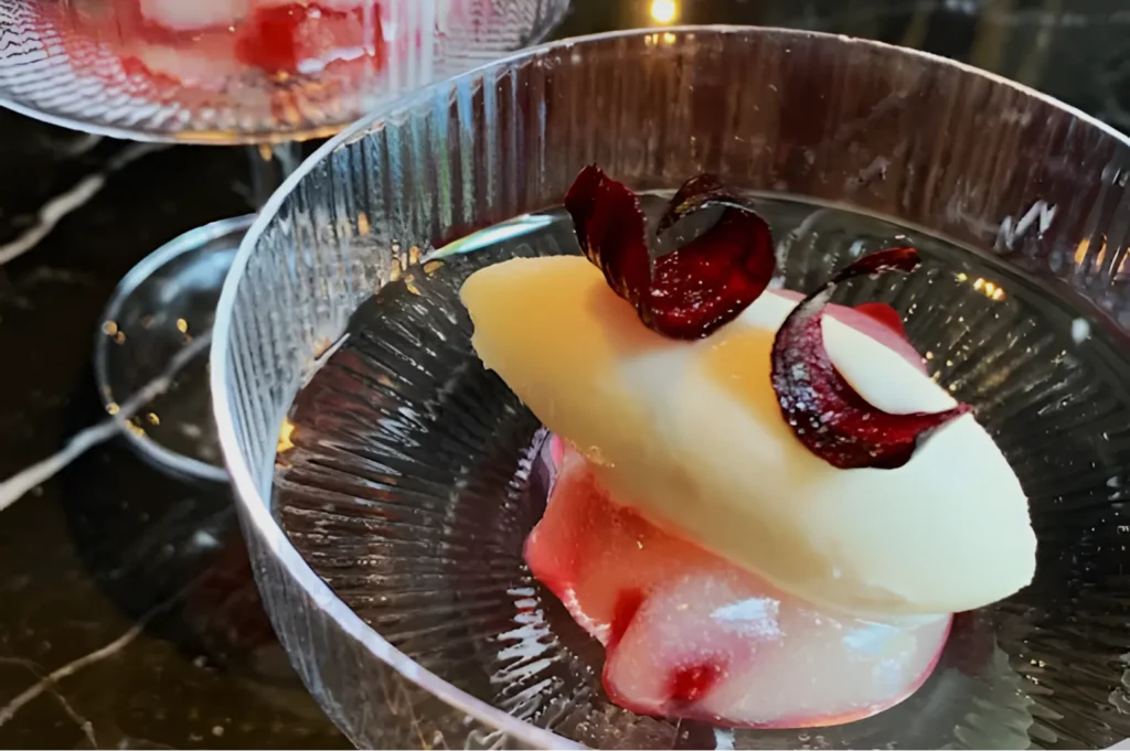 A clear glass dish showcases a creamy white dessert garnished with delicate red curls and a pink sauce beneath, set on a dark marble surface.