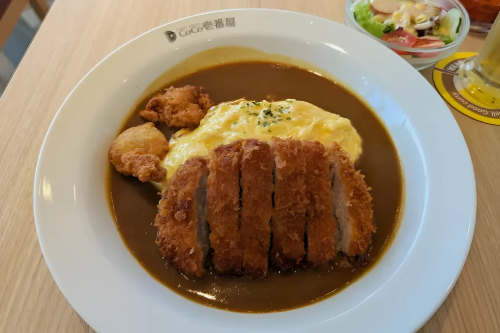 Plate of Japanese curry with golden breaded pork cutlet, fluffy omelette, and fried chicken, garnished with herbs. Served with a side salad.