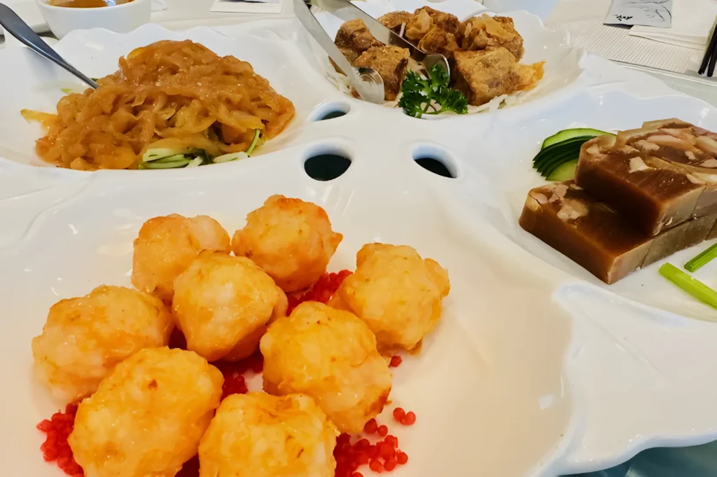 A white platter features golden shrimp balls, jellyfish salad, crispy rolls, and sliced aspic. The dish is colorful and elegantly presented.
