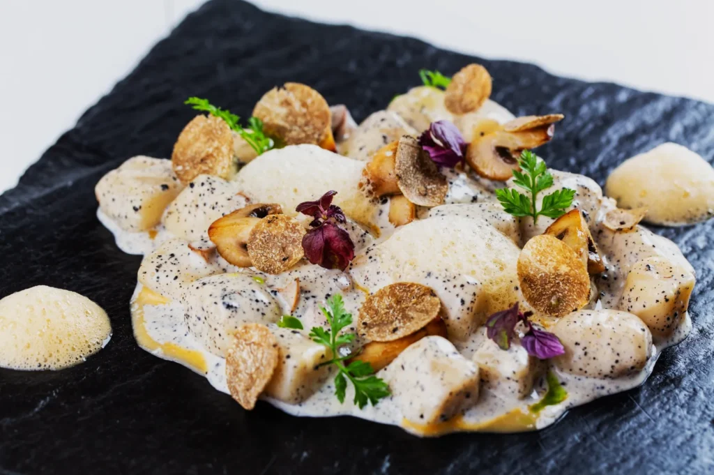 Elegant dish of gnocchi in creamy sauce with truffle slices, mushrooms, and foam on a dark slate plate. Garnished with herbs and purple leaves.