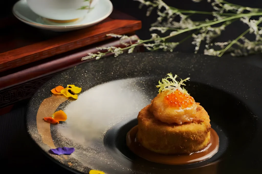 A gourmet dish on a black plate features a round fried cake topped with orange caviar and decorative greens, surrounded by vibrant flower petals.