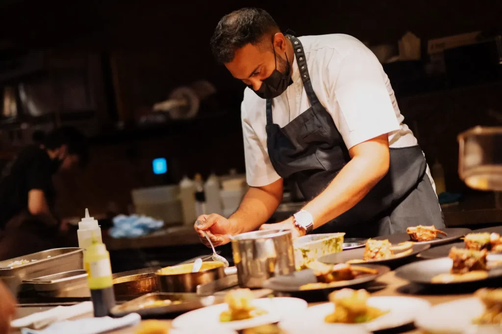 Chef in a kitchen, wearing a mask and apron, artfully plating dishes with focused attention. Warm lighting creates a professional, dedicated ambiance.