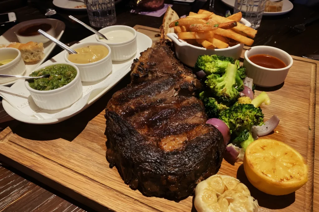 A grilled steak on a wooden board with fries, broccoli, and sliced lemon. Surrounding sauces and roasted garlic create a rich, hearty meal ambiance.