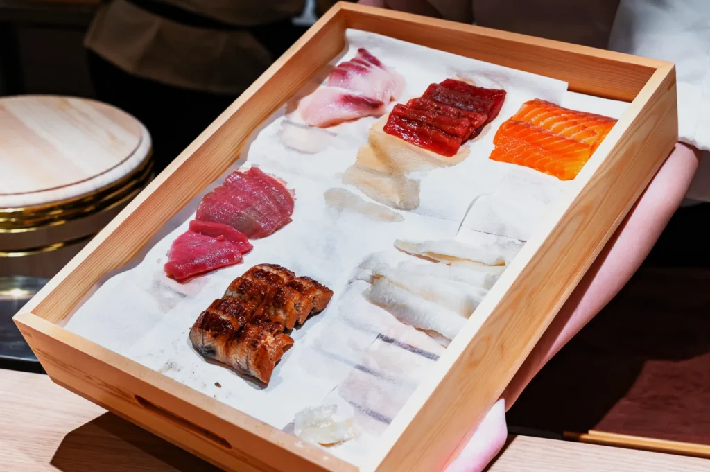 A wooden tray holds various fresh sashimi slices, such as salmon, tuna, and eel, neatly arranged on a white cloth, conveying a sense of freshness and artistry.