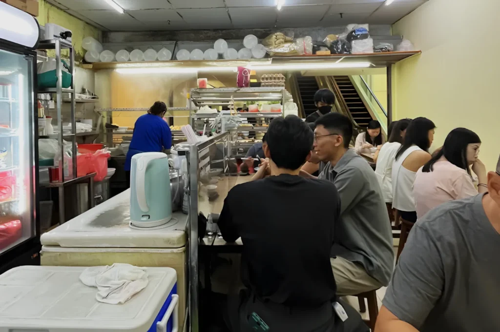 Interior of a busy casual eatery with people seated at a narrow counter. A worker in blue prepares food. Bright lighting, informal atmosphere.